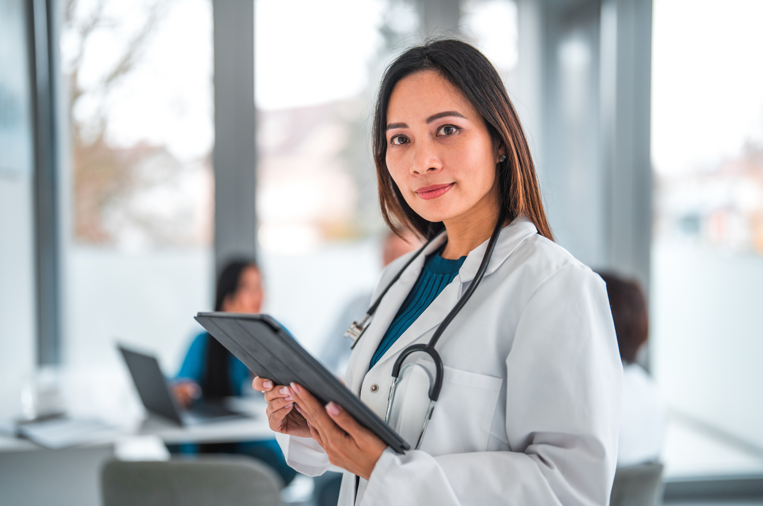 A confident female Plastic Surgeon holds a tablet and looks into the camera.
